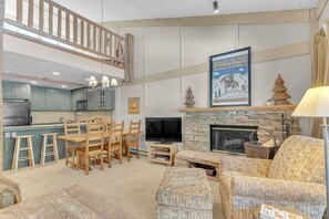 A spacious living room with a high ceiling, wooden furniture, a fireplace, and a mounted television.