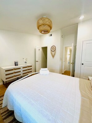 A bedroom with a large bed, a white dresser with drawers, a hanging dreamcatcher, and a ceiling light fixture.