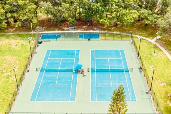 A tennis court with two courts, surrounded by a fence, trees, and a bench.