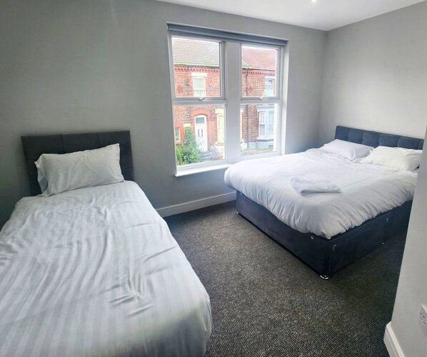 A hotel room with two single beds, a large window with a view of a red brick building, and a carpeted floor. - The Red Lounge Rooms Near Anfield (Liverpool)