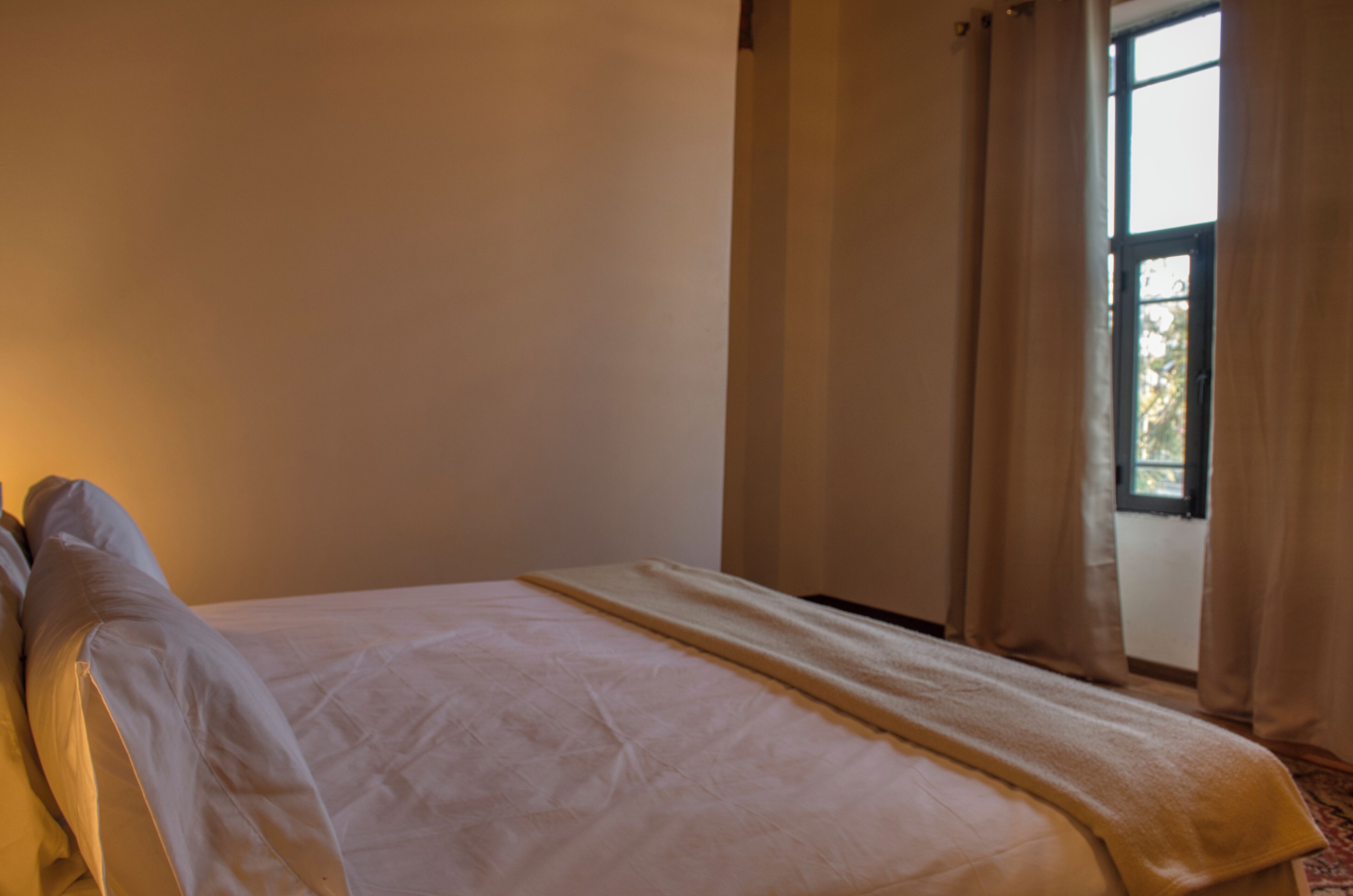 Un lit bien fait, avec une couette blanche et un couvre-lit beige, une fenêtre avec des rideaux et un mur sobre.