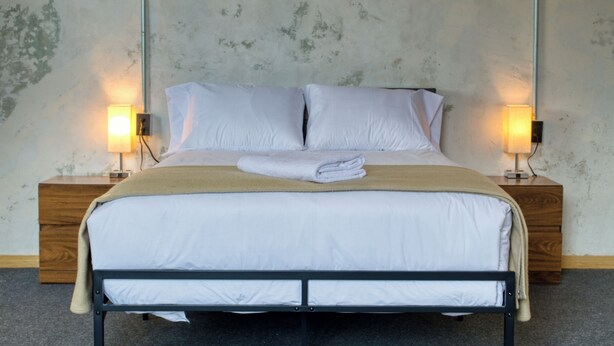 Una cama bien hecha con sábanas blancas y una manta color beige, flanqueada por dos mesitas de noche con lámparas, en una habitación con pared texturizada.