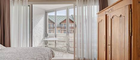 A bedroom with a bed, a wooden wardrobe, a balcony with a table and chairs, and a view of buildings.