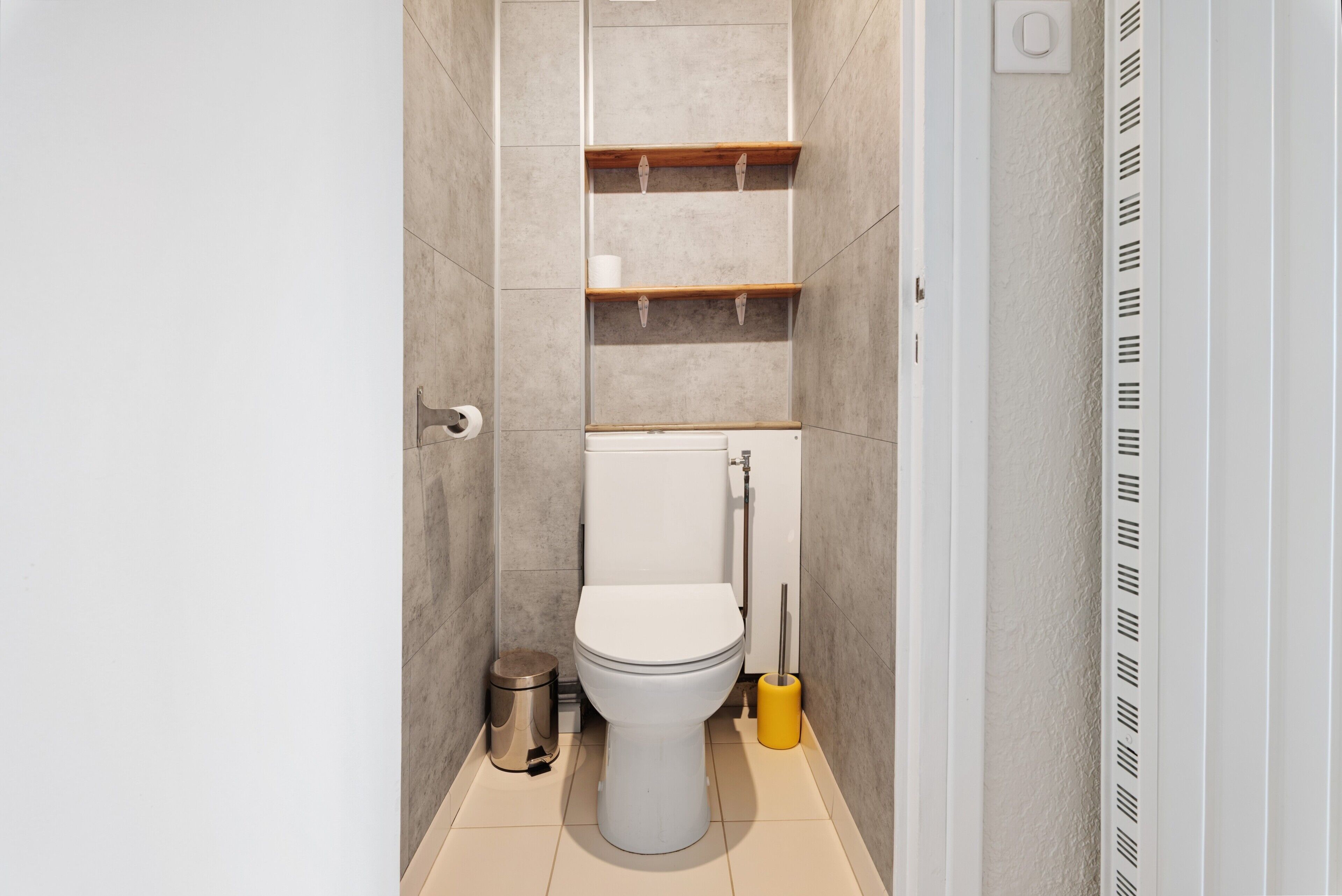 Un baño con inodoro, un cubo de basura, un cepillo para el inodoro y un estante con artículos de aseo.