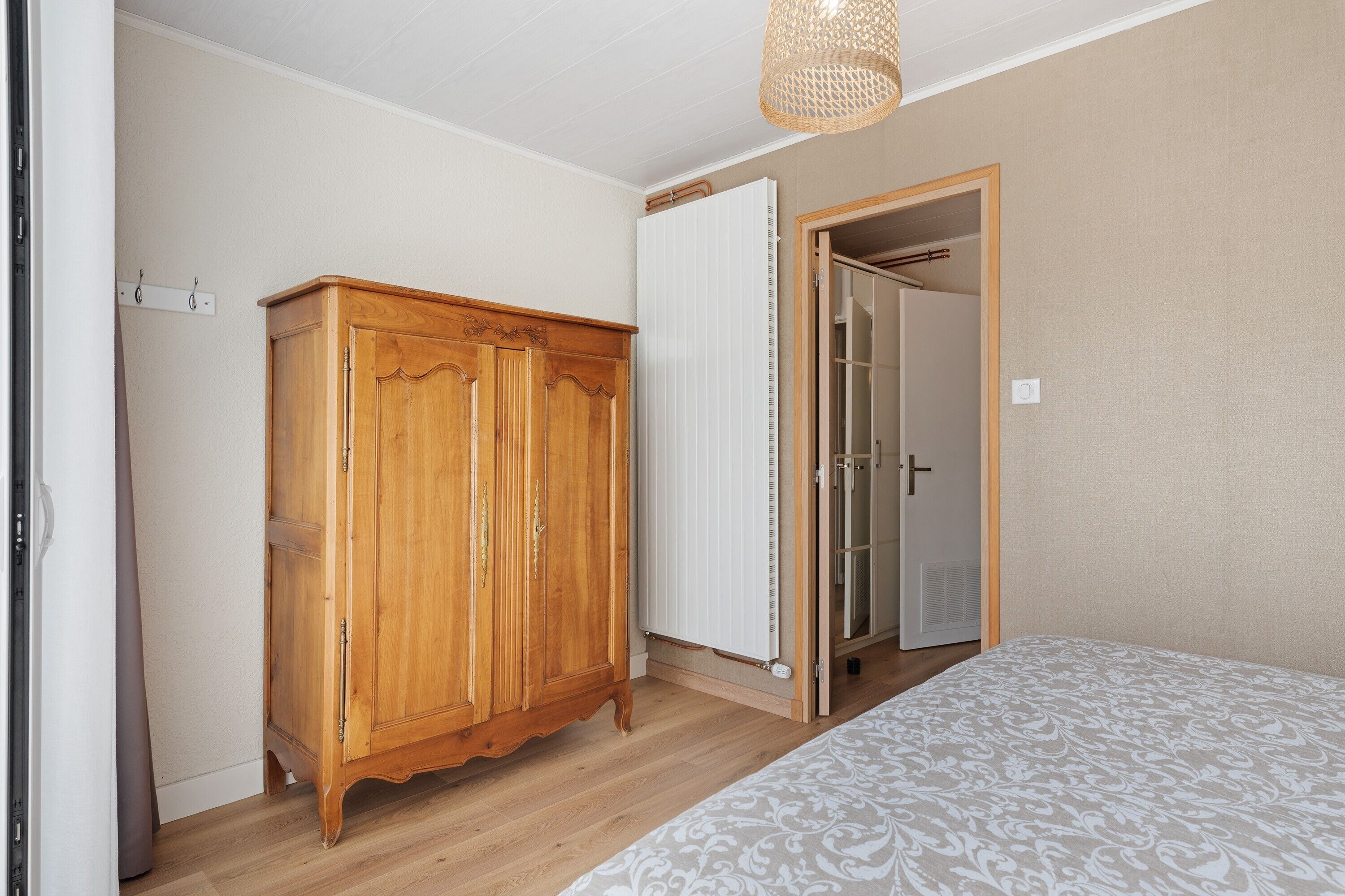 Quarto com guarda-roupa de madeira, radiador, cama com roupa de cama estampada e porta de entrada para um banheiro.