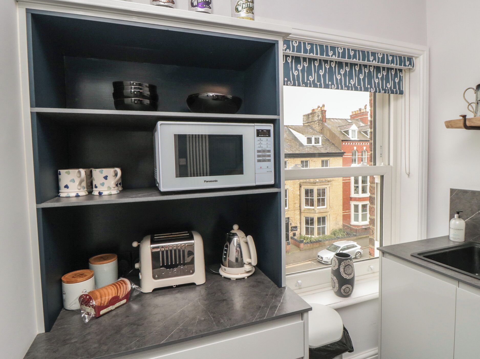 A kitchen with a microwave, toaster, and tea bags on shelves, a window with a view of buildings, and a sink with a bottle.