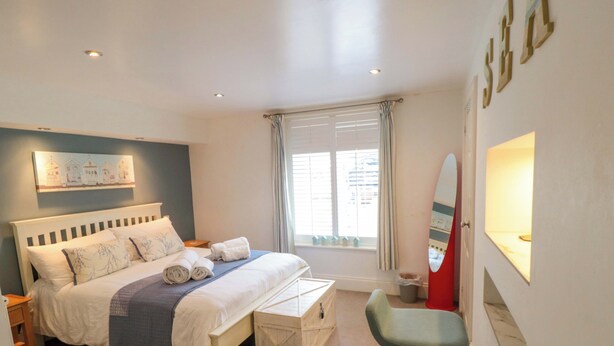 A bedroom with a bed, a bedside table, a chair, a window with curtains, and a wall-mounted mirror. - The Hideaway (Whitby)