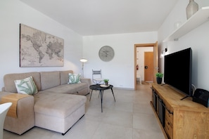 Een woonkamer met een hoekbank, een ronde tafel met een plant, een wereldkaart aan de muur, een klok en een televisie op een houten dressoir.