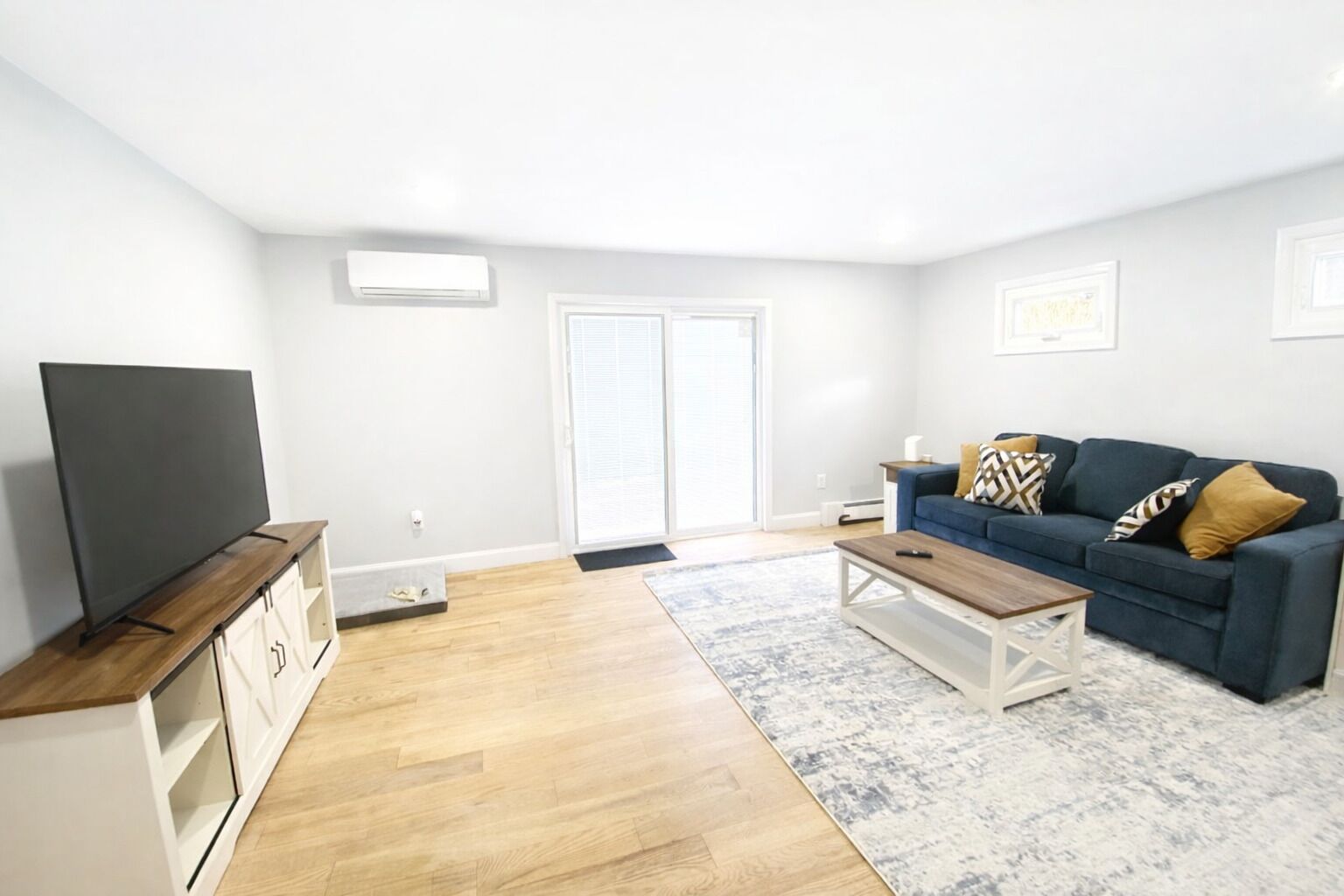 A living room with a flat-screen TV, a blue sofa with patterned cushions, a wooden coffee table, and a large window with blinds.