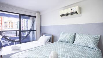 A hotel room with a bed, a window with a view of buildings, an air conditioning unit, and a hammock.