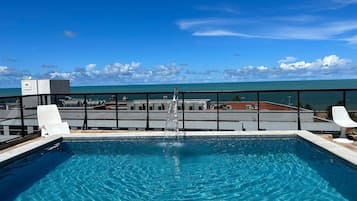 A rooftop pool with clear blue water, a water fountain, and a view of the ocean and buildings in the background.