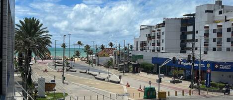 A street view with a beach in the background, palm trees, and a building with a blue awning.