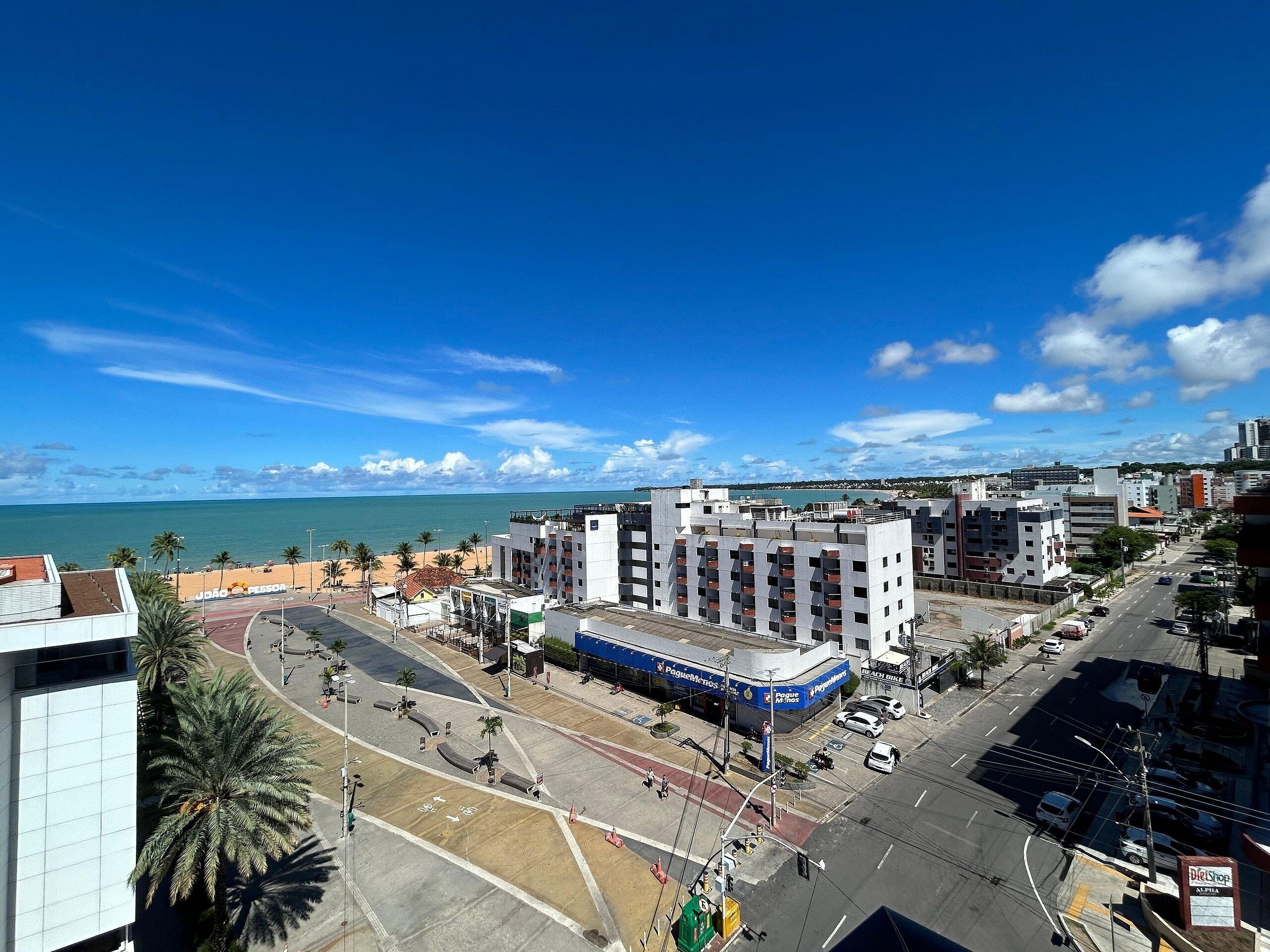 A coastal cityscape with a prominent building featuring a blue sign, a road with a bike lane, and a clear view of the sea.