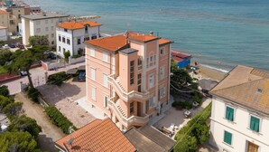 Un bâtiment situé sur la côte, doté d’un balcon, et donnant sur une plage où se trouvent des personnes. On aperçoit également un voilier au loin.