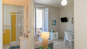 Une salle de bain avec douche, toilettes et lavabo ; un espace de vie avec un canapé, une table et une lampe ; et une chambre à coucher avec un lit et un bureau.