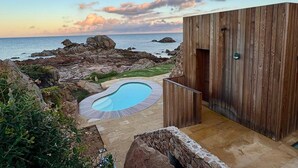 Een moderne buitenzwembadzone met een houten terras, een pad van natuursteen en uitzicht op de oceaan en de rotsachtige kustlijn.