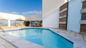 Una piscina con sillas de descanso y sombrillas, rodeada por un edificio blanco con listones de madera.