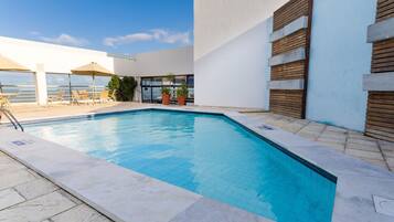 Una piscina con sillas de descanso y sombrillas, rodeada por un edificio blanco con listones de madera.