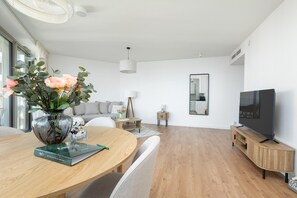 Una sala de estar moderna con comedor, un conjunto de sofá, una mesa de centro, un jarrón con flores, una lámpara, un televisor sobre un mueble de madera y un espejo grande en la pared.