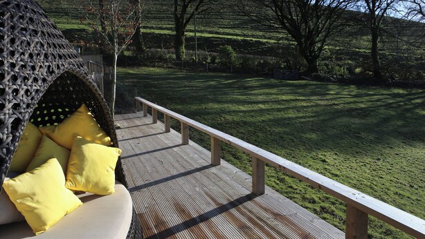 Een houten terras met een witte bank en gele kussens, met uitzicht op een grasachtig landschap met bomen en heuvels.