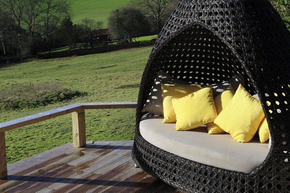 Ein Sitzbereich im Freien aus Weidengeflecht mit Kissen, einer Holzterrasse und Blick auf eine Wiese.