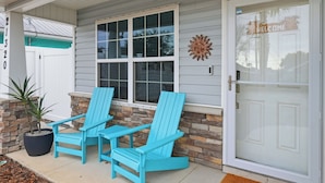Una casa con un porche, dos sillas Adirondack azules, un muro de piedra y una puerta blanca con un letrero que dice 'WELCOME'.