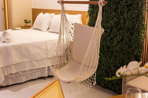 Una cama con sábanas blancas, un sillón colgante de macramé y una pared cubierta de follaje verde.