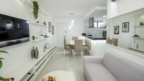 Uma sala de estar moderna com televisão de tela plana, um sofá, uma área de jantar com mesa e cadeiras, e uma cozinha ao fundo.
