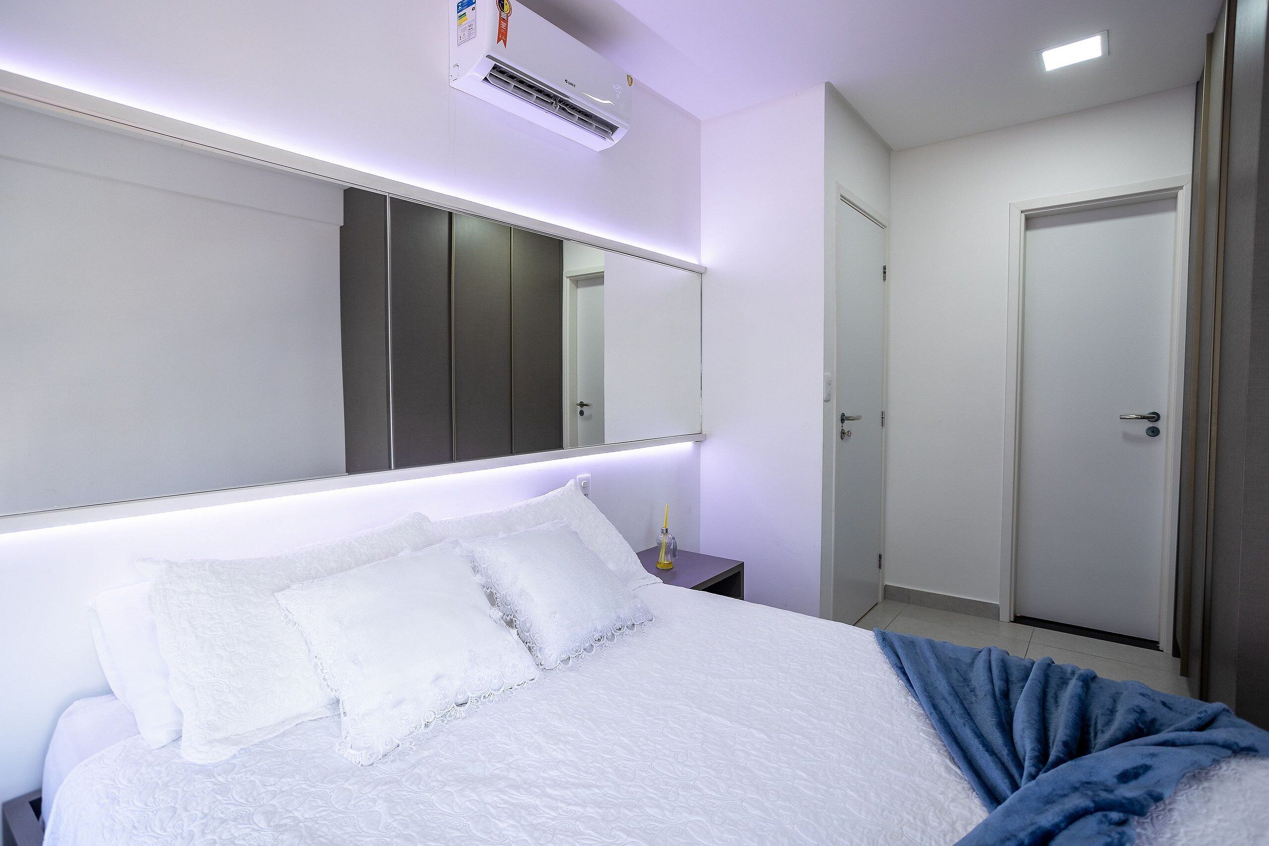 A hotel room with a large bed, a bedside table, a mirror, an air conditioning unit, and a door.