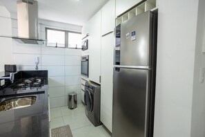 A compact kitchen with a stainless steel refrigerator, a washing machine, a stove, and a window above the oven. - Tropical Hub #1002 - Apartment in Manaira by Carpediem (João Pessoa)