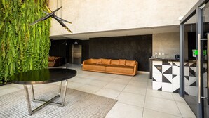 Um hall com sofá, mesa, tapete estampado, parede verde com plantas e balcão xadrez.