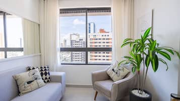 Un salon avec un canapé, un fauteuil, une plante en pot et une vue sur la ville par la fenêtre.