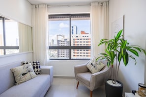 A living room with a sofa, armchair, potted plant, and a city view through a window. - Tropical Hub #1002 - Apartment in Manaira by Carpediem (João Pessoa)