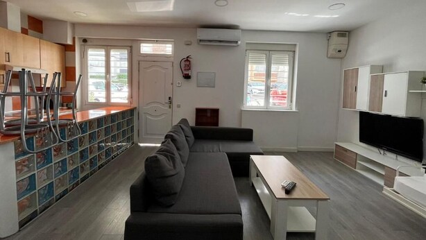 Uma sala de estar moderna com sofá, mesa de centro e televisão. Há uma área de cozinha com armários e bancada.
