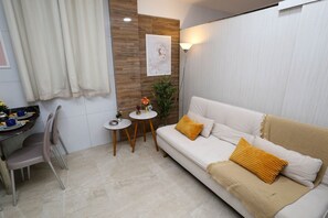 Uma sala de estar moderna com um sofá bege, duas almofadas amarelas, uma mesa redonda pequena com uma planta, uma cadeira e uma obra de arte pendurada na parede.