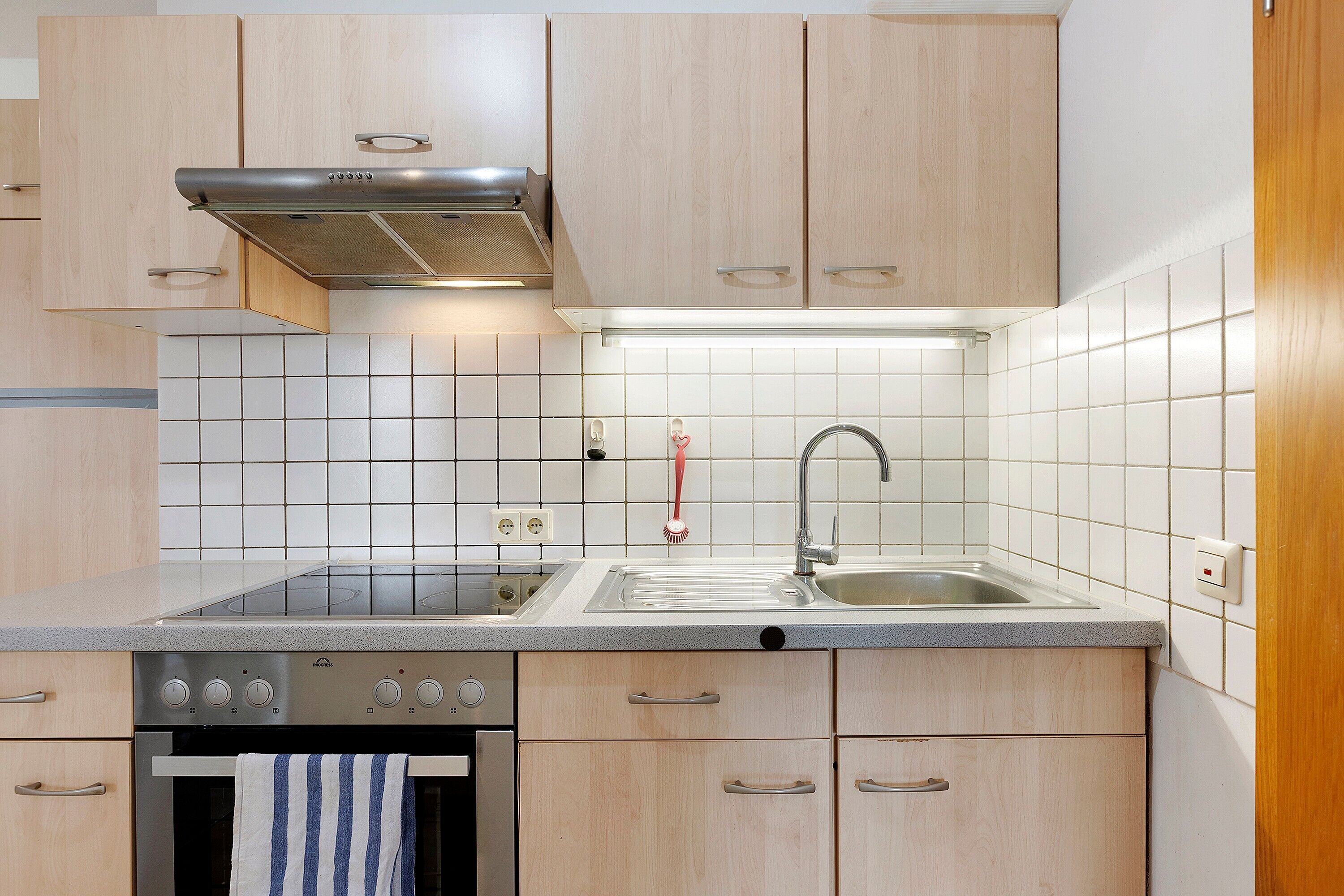 A kitchen with light wood cabinets, a stainless steel range hood, a double sink, and a built-in oven.