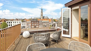 A wooden deck with wicker chairs and a table, overlooking a cityscape with historic buildings and a tower. - Hotel Copper (Middelburg)