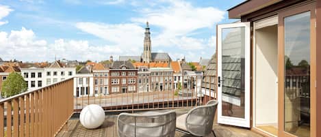 A wooden deck with wicker chairs and a table, overlooking a cityscape with historic buildings and a tower.