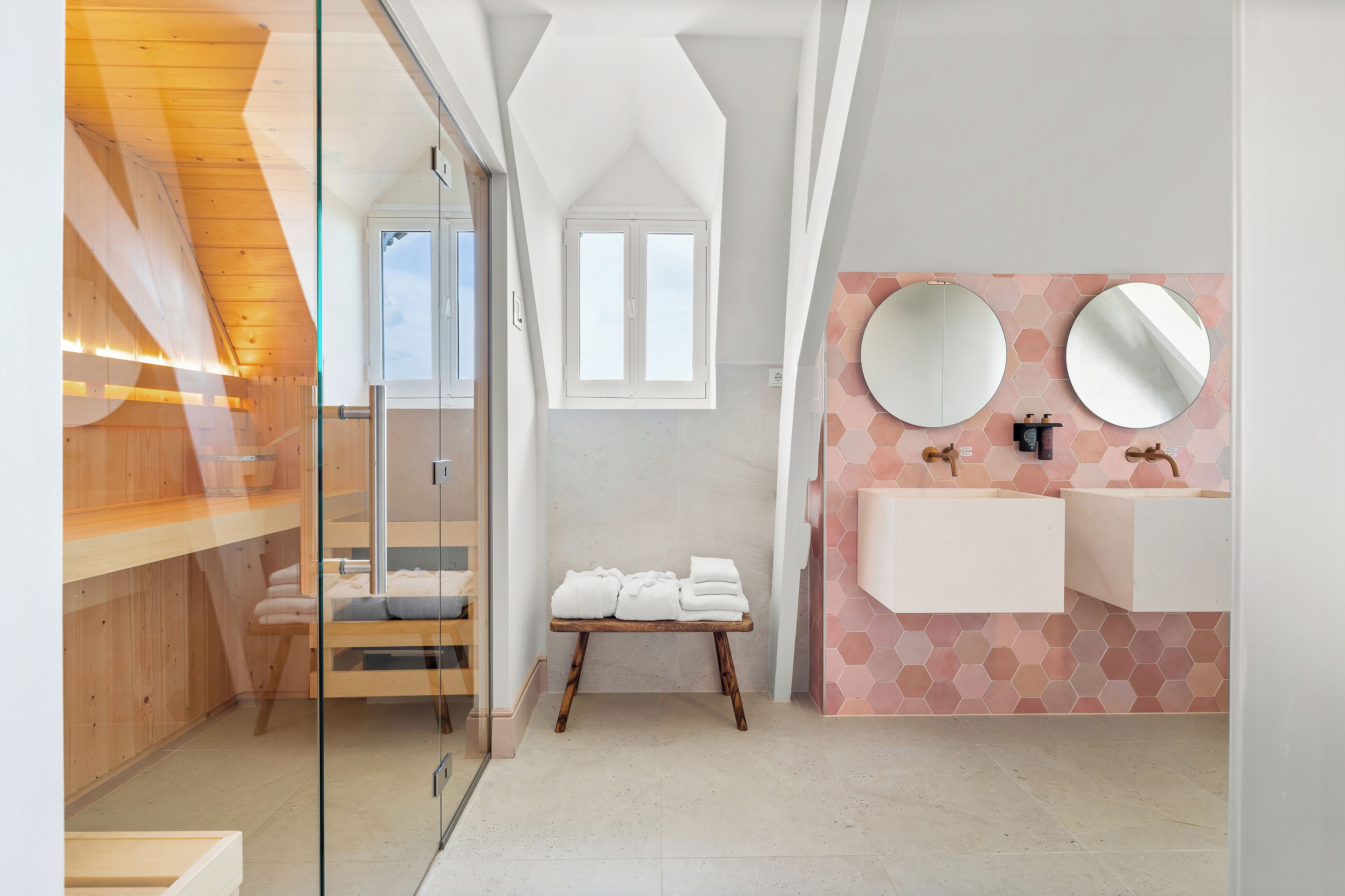 A modern bathroom with a glass shower enclosure, wooden benches, a small table with towels, and a pink hexagonal tile backsplash.