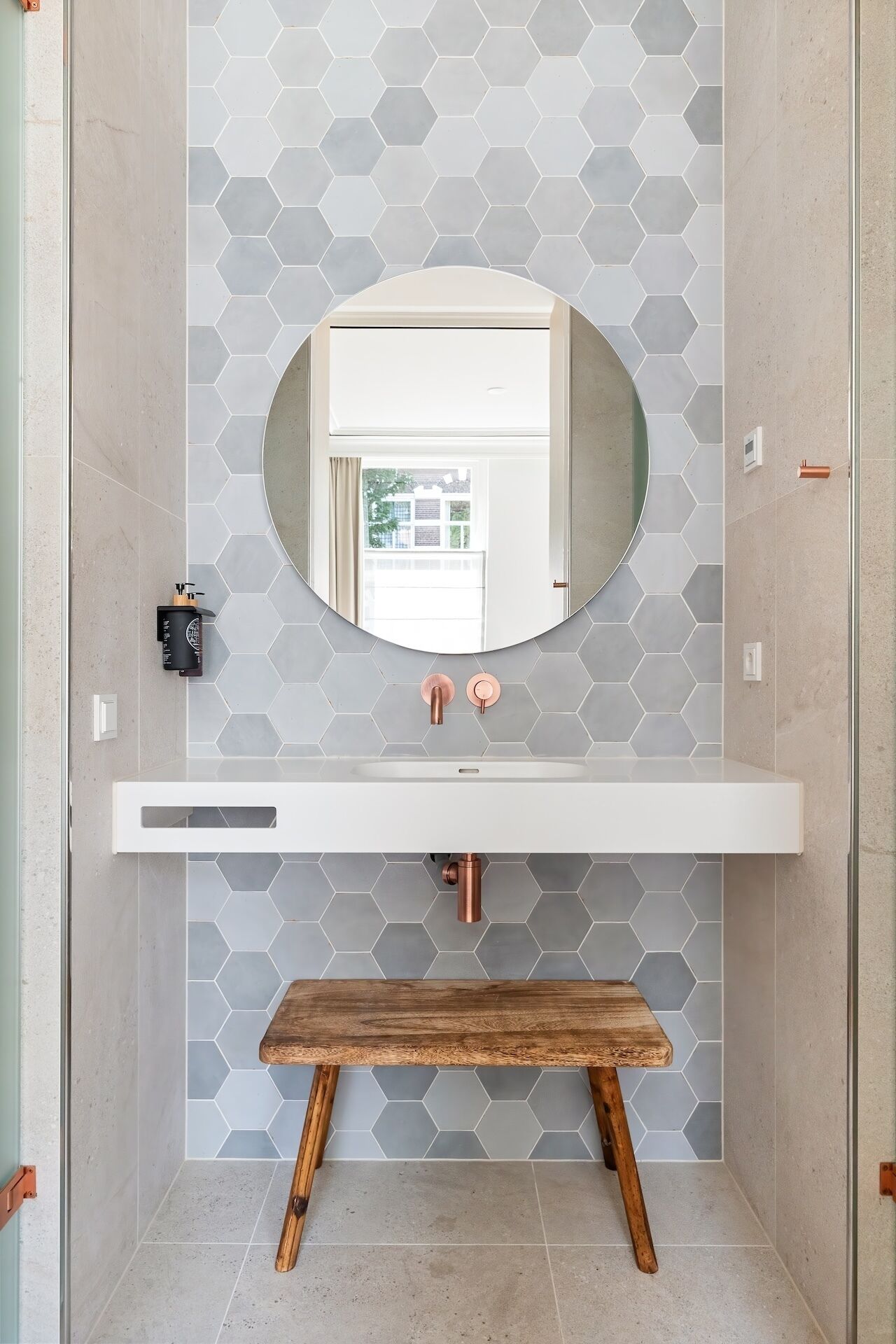 Un baño con un salpicadero de azulejos hexagonales, un lavamanos blanco con accesorios de cobre y un banco de madera.