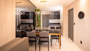 A modern dining area with a rectangular table set for six, surrounded by chairs. A kitchen is visible in the background with a clock on the wall. - Mindset #207 - Comfort and Location by Carpediem (João Pessoa)