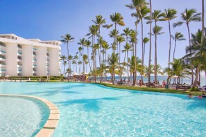Área de piscina em resort, com piscina azul límpida, palmeiras e um prédio de hotel ao fundo.