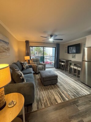A living room with a sofa, ottoman, and a dining area with bar stools. - Myrtle Beach Please! Newly Remodeled Apartment Awaits. (Myrtle Beach)