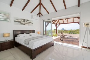 A bedroom with a large bed, a nightstand, a lamp, and a view of a balcony with a chair. - Villa Joahia - Three Bedroom Villa, Sleeps 7 (Le Diamant)