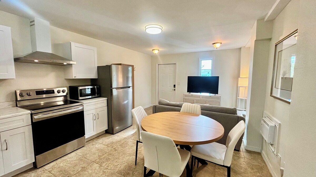 A compact kitchen with a stove, microwave, and refrigerator, a dining table with chairs, and a television in the background.