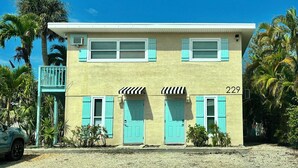 A two-story building with a balcony, numbered 229, featuring a light yellow facade, turquoise shutters, and a small awning over the doors. - SunShell 2 - Steps from Sand (Fort Myers)