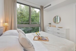 Een slaapkamer met een groot raam, een nachtkastje met een lamp, een kaptafel en een dienblad met gebakjes en bloemen op het bed.
