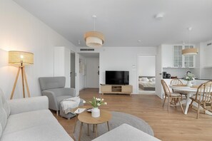 Een moderne woonkamer met een grijze bank, een salontafel en een televisie. Er is een zithoek met een witte tafel en stoelen.