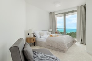A bedroom with a bed, bedside tables, a lamp, a view of the sea, and a window with curtains.