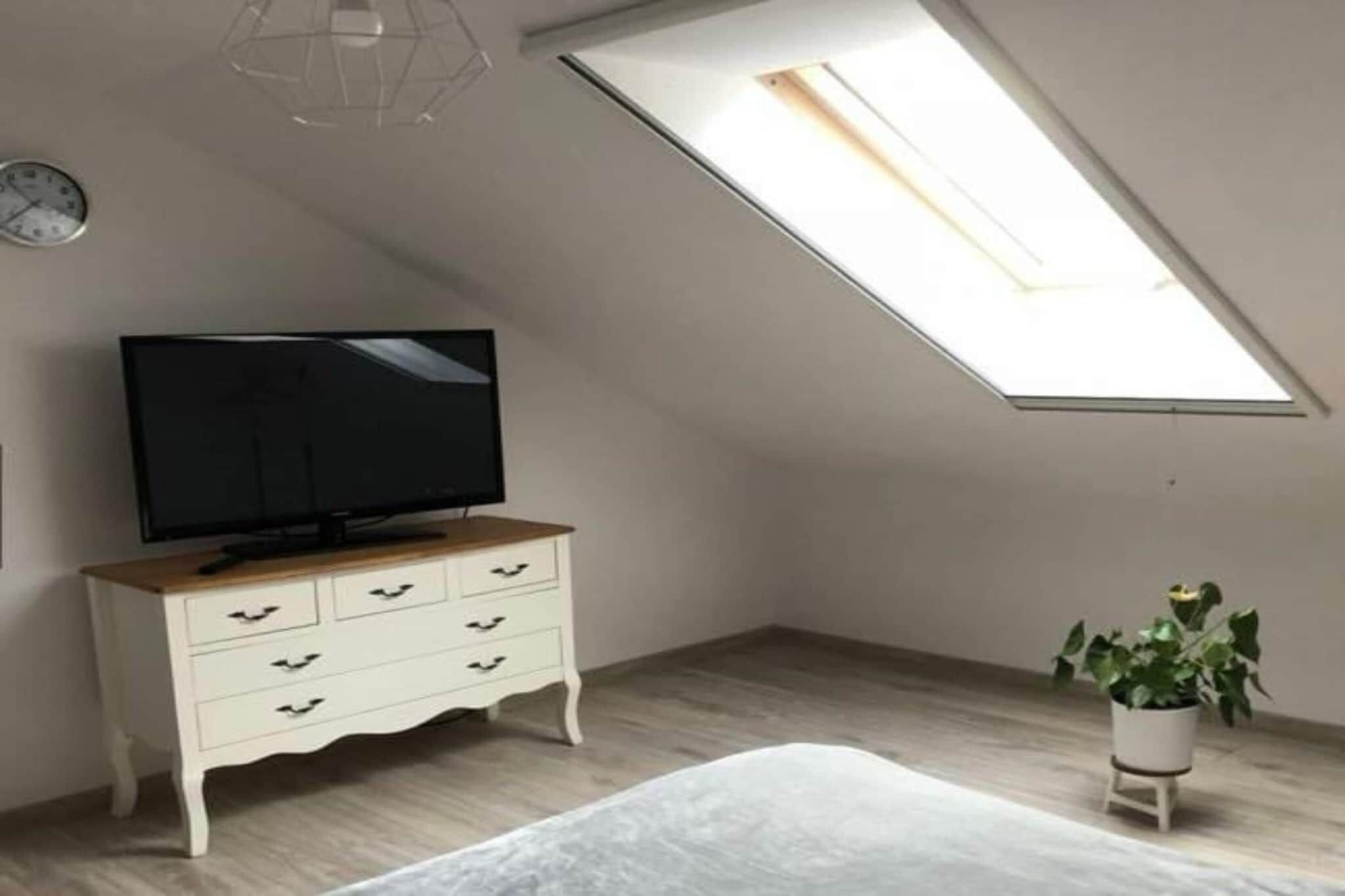 Une chambre avec un plafond en pente, un téléviseur posé sur une commode, un lit et une plante en pot.
